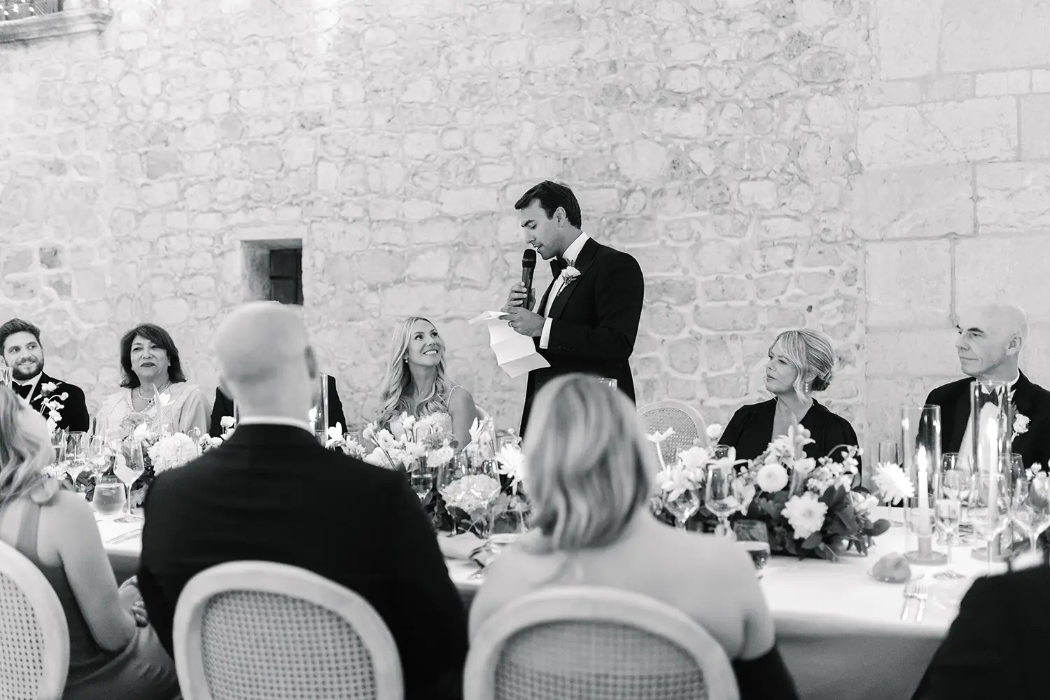 Black-and-white photo of a groom giving a heartfelt speech at an intimate wedding reception. The bride and close guests sit around the table, smiling warmly, reflecting the simplicity and sincerity of smaller celebrations.